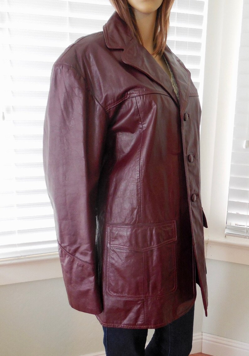 May include: A burgundy leather jacket with a button closure and a single pocket on the front. The jacket has a collar and long sleeves.
