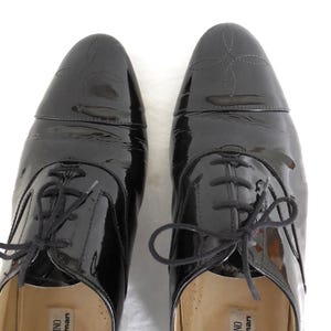 Puede incluir: Un par de zapatos de vestir negros de charol con acabado brillante. Los zapatos tienen un estilo Oxford cl&aacute;sico con cierre de cordones y costuras decorativas en la puntera. La marca "VALENTINO for Jarman" es visible en el interior.