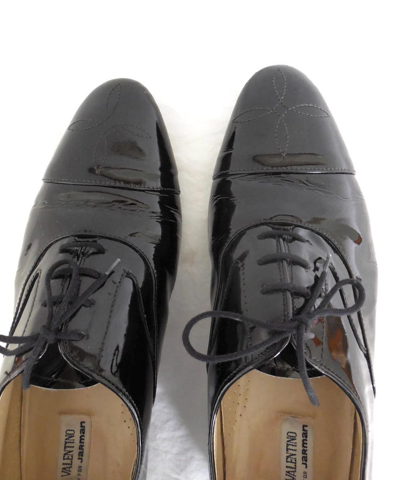 Puede incluir: Un par de zapatos de vestir negros de charol con acabado brillante. Los zapatos tienen un estilo Oxford cl&aacute;sico con cierre de cordones y costuras decorativas en la puntera. La marca "VALENTINO for Jarman" es visible en el interior.