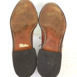 May include: A pair of brown leather dress shoes, viewed from the soles up. The soles show wear and tear, with visible stitching around the edges. The heels are black rubber. The shoes are laced.