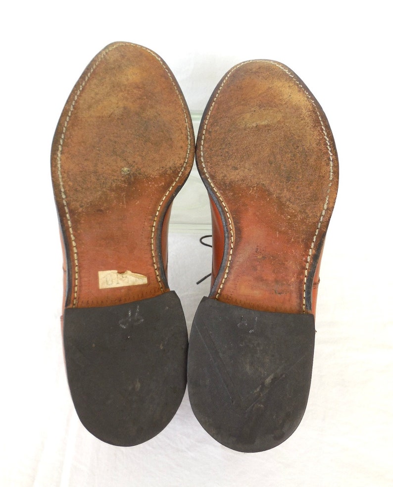 May include: A pair of brown leather dress shoes, viewed from the soles up. The soles show wear and tear, with visible stitching around the edges. The heels are black rubber. The shoes are laced.