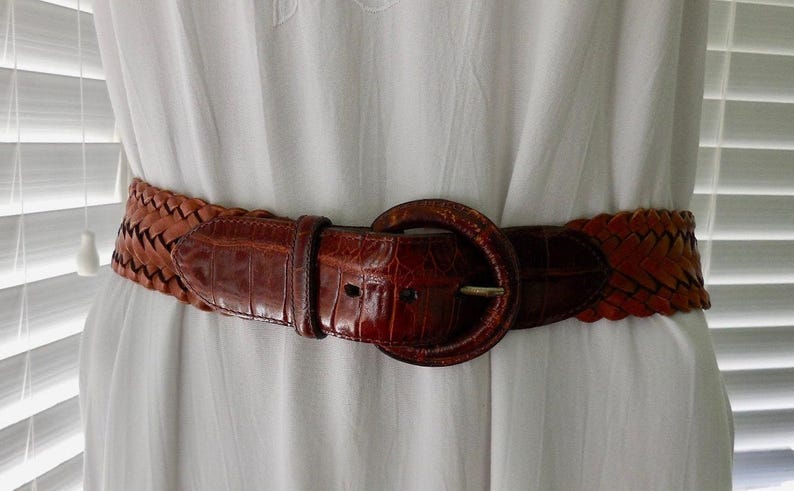 May include: A brown leather belt with a braided design and a large, round buckle. The belt is worn around the waist of a white dress.