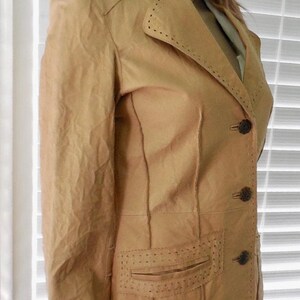 May include: A tan leather jacket with a notched collar, long sleeves, and a single-breasted closure. The jacket has a pocket on the left side and decorative stitching throughout.
