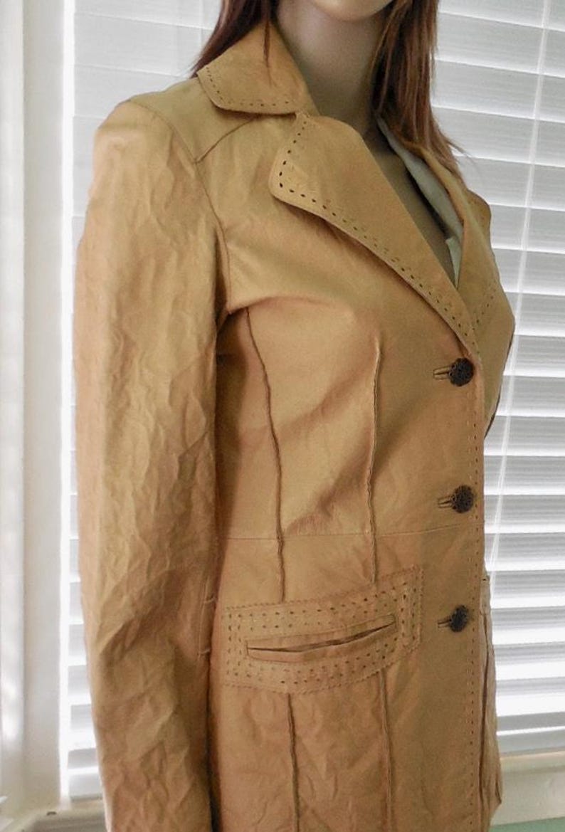 May include: A tan leather jacket with a notched collar, long sleeves, and a single-breasted closure. The jacket has a pocket on the left side and decorative stitching throughout.