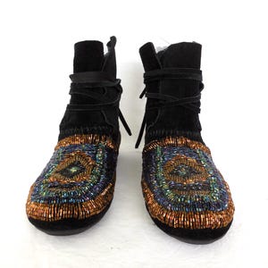K&ouml;nnte beinhalten: Ein Paar schwarze Wildleder-Ankle Boots mit aufwendiger Perlenarbeit. Die Stiefel verf&uuml;gen &uuml;ber einen Wickelverschluss und ein farbenfrohes Muster aus orangefarbenen, blauen und gr&uuml;nen Perlen im Zehenbereich, was ein einzigartiges und stilvolles Design ergibt.