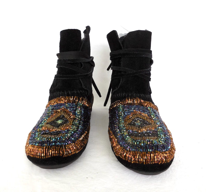 K&ouml;nnte beinhalten: Ein Paar schwarze Wildleder-Ankle Boots mit aufwendiger Perlenarbeit. Die Stiefel verf&uuml;gen &uuml;ber einen Wickelverschluss und ein farbenfrohes Muster aus orangefarbenen, blauen und gr&uuml;nen Perlen im Zehenbereich, was ein einzigartiges und stilvolles Design ergibt.