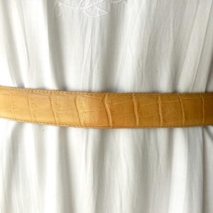 May include: A tan-coloured belt with a crocodile skin pattern. The belt is worn over a white dress. The belt is made of leather and has a rectangular buckle. The belt is approximately 3.8 cm wide.