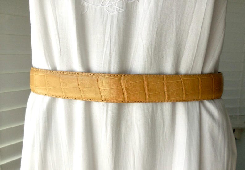 May include: A tan-coloured belt with a crocodile skin pattern. The belt is worn over a white dress. The belt is made of leather and has a rectangular buckle. The belt is approximately 3.8 cm wide.