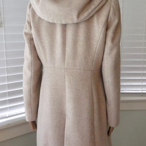 May include: A beige hooded coat with a tailored silhouette. The coat features a high collar, a fitted waist, and a flared skirt. The hood is large and drapes over the shoulders. The coat is made of a soft, wool-like material.