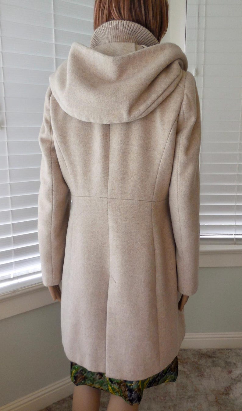 May include: A beige hooded coat with a tailored silhouette. The coat features a high collar, a fitted waist, and a flared skirt. The hood is large and drapes over the shoulders. The coat is made of a soft, wool-like material.