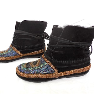 K&ouml;nnte beinhalten: Ein Paar schwarze Wildleder-Ankle-Boots mit Perlenbesatz. Die Stiefel haben einen Wickelverschluss und ein farbenfrohes Perlendesign in Blau, Gr&uuml;n und Gold. Die Sohlen sind schwarz.