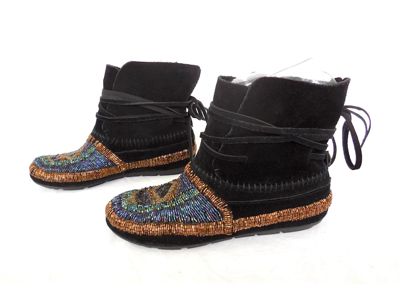 K&ouml;nnte beinhalten: Ein Paar schwarze Wildleder-Ankle-Boots mit Perlenbesatz. Die Stiefel haben einen Wickelverschluss und ein farbenfrohes Perlendesign in Blau, Gr&uuml;n und Gold. Die Sohlen sind schwarz.