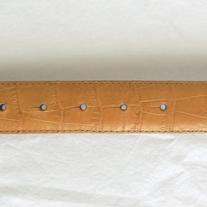 May include: Tan belt with a crocodile skin effect. The belt has five holes and a rounded end. The stitching is visible along the edges. The belt is made of a durable material. The belt's length is approximately 100 cm.