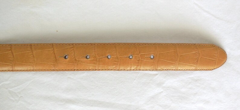 May include: Tan belt with a crocodile skin effect. The belt has five holes and a rounded end. The stitching is visible along the edges. The belt is made of a durable material. The belt's length is approximately 100 cm.