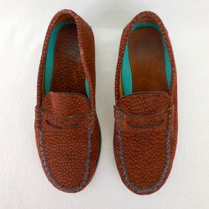 May include: A pair of brown loafers featuring a textured, pebbled leather exterior with a classic penny slot strap across the vamp. The interior lining is a vibrant turquoise color, and the shoes have visible stitching along the rounded toe box.
