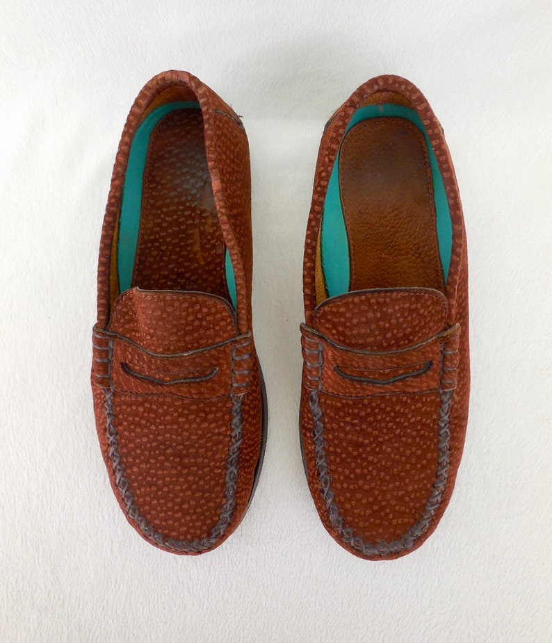 May include: A pair of brown loafers featuring a textured, pebbled leather exterior with a classic penny slot strap across the vamp. The interior lining is a vibrant turquoise color, and the shoes have visible stitching along the rounded toe box.
