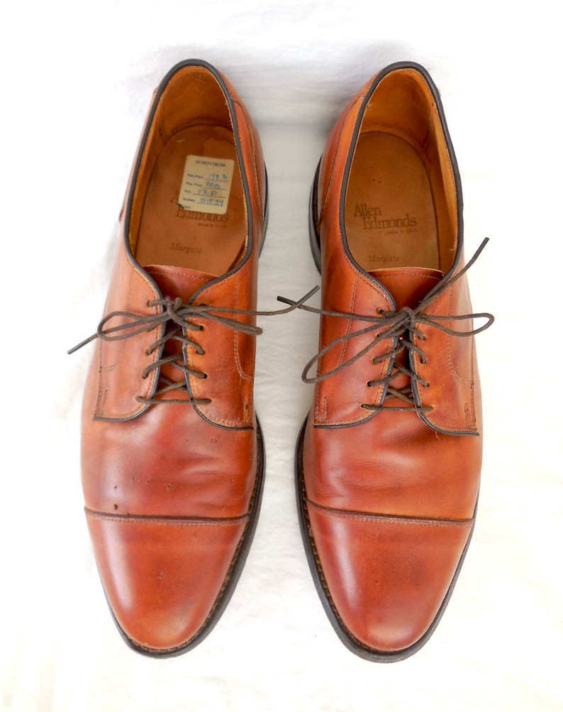 May include: A pair of brown leather Allen Edmonds Margate dress shoes, viewed from above. The shoes have a classic cap-toe design with brown laces and a dark brown sole. The interior of the shoe is tan.