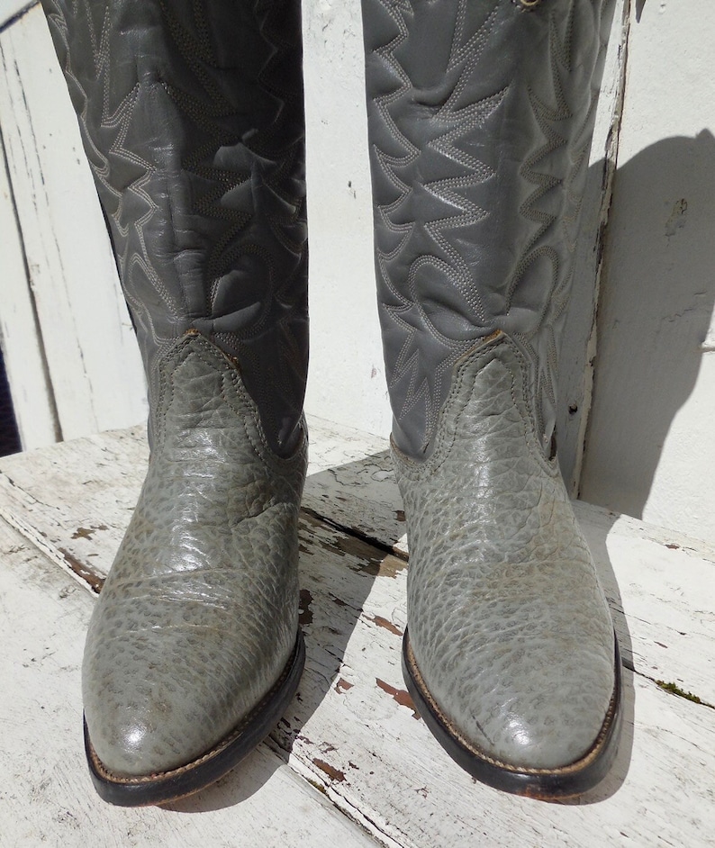 May include: A pair of gray cowboy boots. The boots have a textured, almost reptilian appearance on the foot and a stitched design on the shaft. The soles are dark brown with a light brown welt. The boots are made of leather.