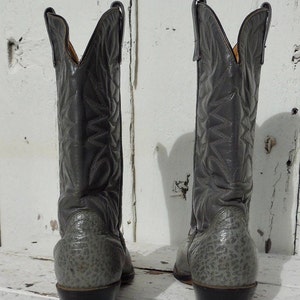 May include: A pair of gray cowboy boots with intricate stitched patterns on the shafts. The boots have a textured design on the lower portion and a classic pointed toe. The heels are dark brown.