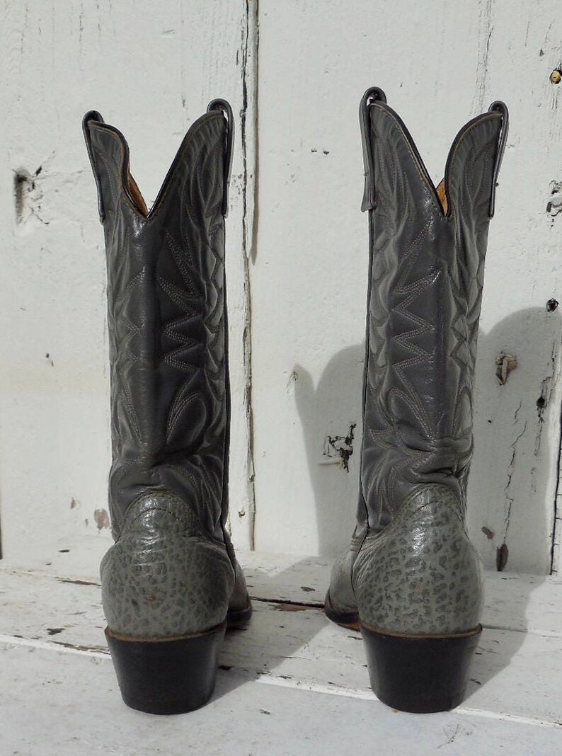 May include: A pair of gray cowboy boots with intricate stitched patterns on the shafts. The boots have a textured design on the lower portion and a classic pointed toe. The heels are dark brown.