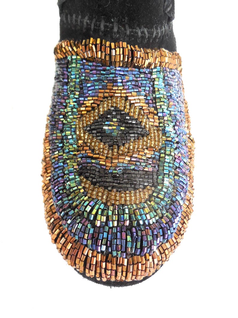 K&ouml;nnte beinhalten: Nahaufnahme einer schwarzen, mit Perlen besetzten Mokassinspitze. Die Spitze ist mit einem Muster aus schillernden Perlen in Gold-, Blau-, Gr&uuml;n- und Lilat&ouml;nen bedeckt. Die Oberseite des Mokassins ist aus schwarzem Wildleder.