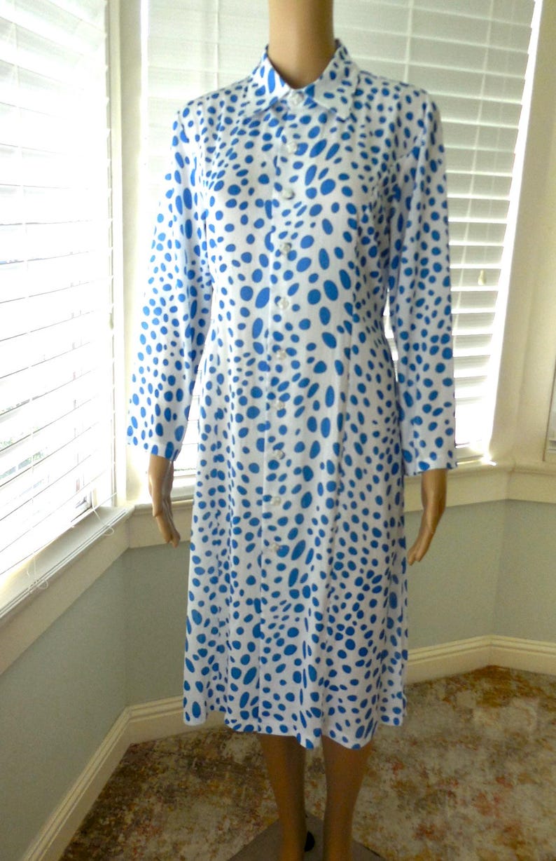 May include: A long-sleeved dress with a white base and a pattern of blue spots. The dress features a collared neckline and buttons down the front. The dress is knee-length, approximately 100 cm.
