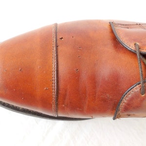 May include: Close-up of a brown leather dress shoe. The shoe features a cap toe design with visible stitching and dark brown laces. The leather has a polished finish and a few minor imperfections.