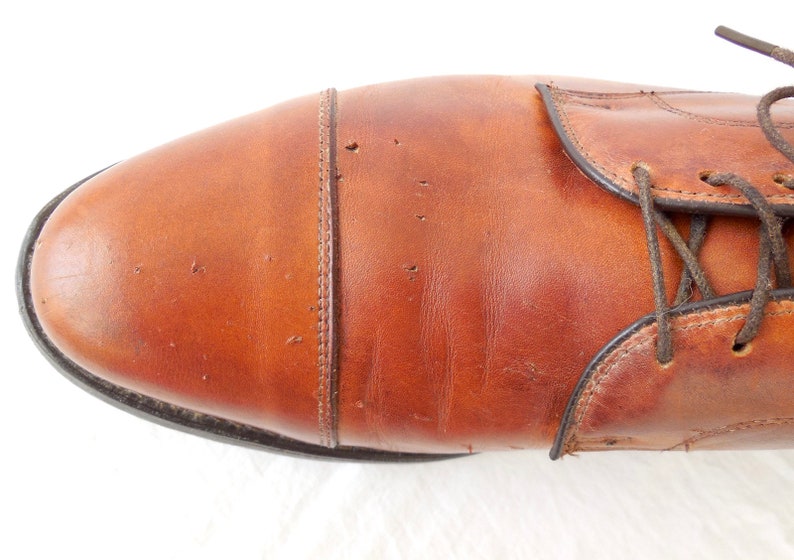 May include: Close-up of a brown leather dress shoe. The shoe features a cap toe design with visible stitching and dark brown laces. The leather has a polished finish and a few minor imperfections.