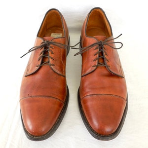 May include: A pair of brown leather dress shoes with dark brown laces. The shoes have a classic cap-toe design and a dark sole. The leather shows signs of wear, giving them a vintage look. The shoes are displayed on a white background.