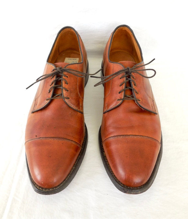 May include: A pair of brown leather dress shoes with dark brown laces. The shoes have a classic cap-toe design and a dark sole. The leather shows signs of wear, giving them a vintage look. The shoes are displayed on a white background.
