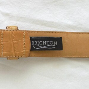 May include: Tan leather belt with a decorative silver-tone buckle. The belt has a textured, crocodile-like pattern and a black tag that reads "BRIGHTON". The belt is a size M.
