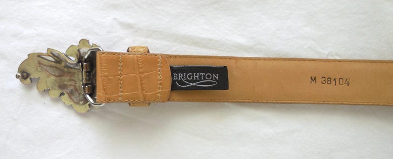 May include: Tan leather belt with a decorative silver-tone buckle. The belt has a textured, crocodile-like pattern and a black tag that reads "BRIGHTON". The belt is a size M.