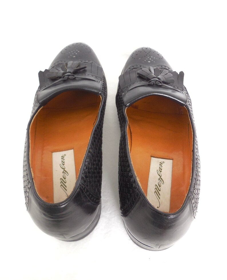 May include: A pair of black leather loafers with a woven pattern and tassel detail. The loafers have a brown leather insole with the brand name "Meglan" printed on them.