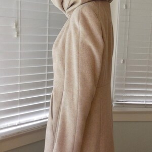 May include: A beige hooded coat with a tailored silhouette. The coat features a large hood, long sleeves, and a knee-length hem. The coat is made of a soft, textured fabric and is shown in profile.