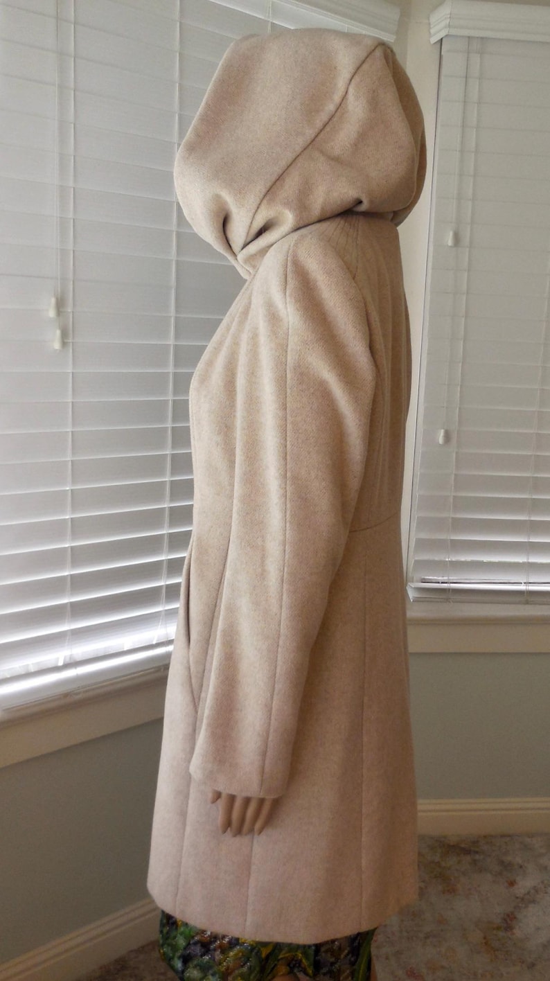 May include: A beige hooded coat with a tailored silhouette. The coat features a large hood, long sleeves, and a knee-length hem. The coat is made of a soft, textured fabric and is shown in profile.