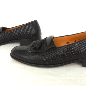 May include: A pair of black leather tassel loafers with a woven pattern. The loafers have a low heel and a pointed toe. The text "84657 404 M" is visible on the insole.