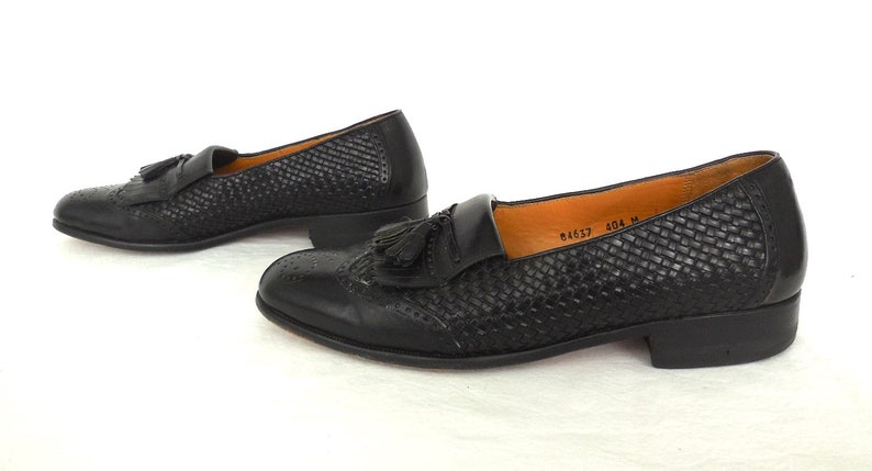 May include: A pair of black leather tassel loafers with a woven pattern. The loafers have a low heel and a pointed toe. The text "84657 404 M" is visible on the insole.