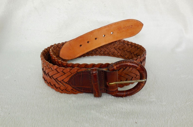 May include: A brown leather belt with a braided design and a round metal buckle. The belt has the number "30 42107" stamped on the back.