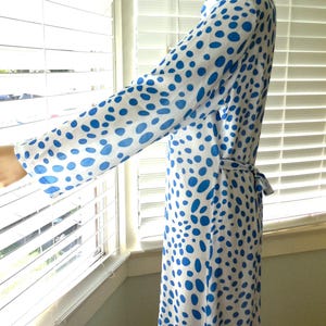 May include: A long, flowing robe in white fabric with a pattern of blue polka dots. The robe has long sleeves and a matching belt. The garment is shown near a window with blinds.