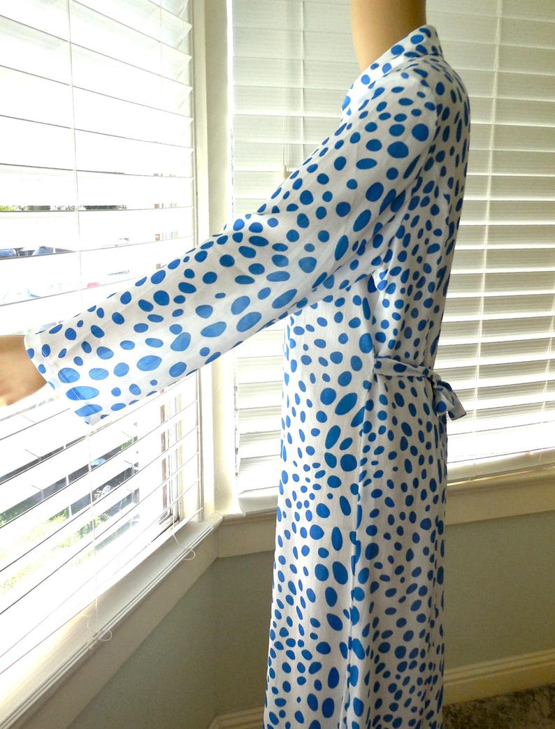May include: A long, flowing robe in white fabric with a pattern of blue polka dots. The robe has long sleeves and a matching belt. The garment is shown near a window with blinds.