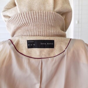 May include: Beige coat with a hood and a ribbed knit collar. The interior lining is a light beige color with a burgundy trim. The label reads "ZARA BASIC".