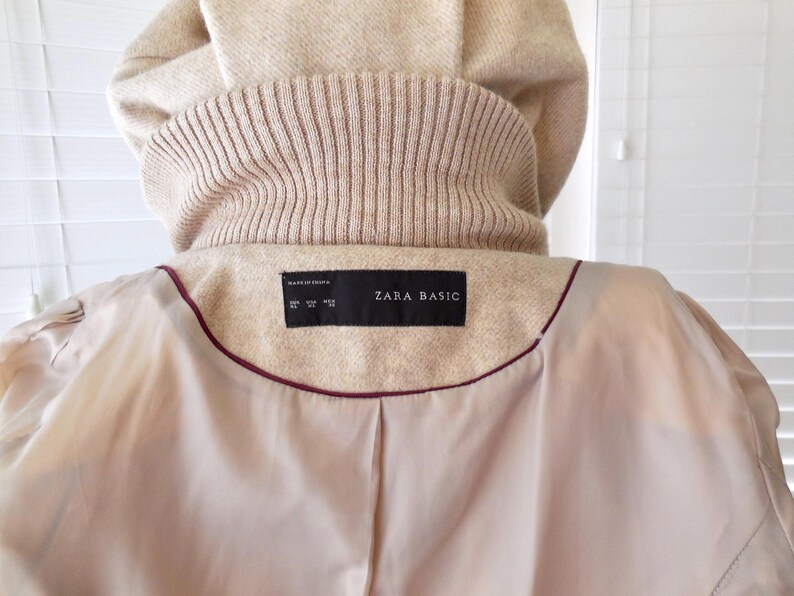 May include: Beige coat with a hood and a ribbed knit collar. The interior lining is a light beige color with a burgundy trim. The label reads "ZARA BASIC".