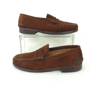 May include: A pair of brown loafers featuring a textured, dotted surface pattern. The shoes have a classic slip-on design with a stitched moccasin-style toe seam, a low heel, and a dark brown sole. The exterior material has a distinct pebbled finish.