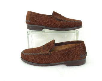 Op de afbeelding: Een paar bruine loafers met een textuur van stippen op het oppervlak. De schoenen hebben een klassiek instapmodel met een gestikte mocassin-naad bij de neus, een lage hak en een donkerbruine zool. Het buitenmateriaal heeft een korrelige afwerking.