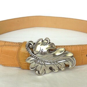 May include: Tan leather belt with a silver-coloured leaf-shaped buckle. The belt has a textured, crocodile-like pattern. The buckle is detailed with a beaded edge and leaf veins.