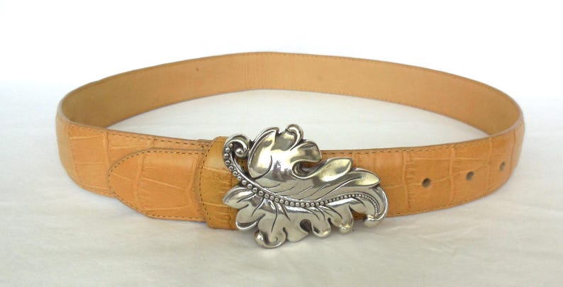 May include: Tan leather belt with a silver-coloured leaf-shaped buckle. The belt has a textured, crocodile-like pattern. The buckle is detailed with a beaded edge and leaf veins.