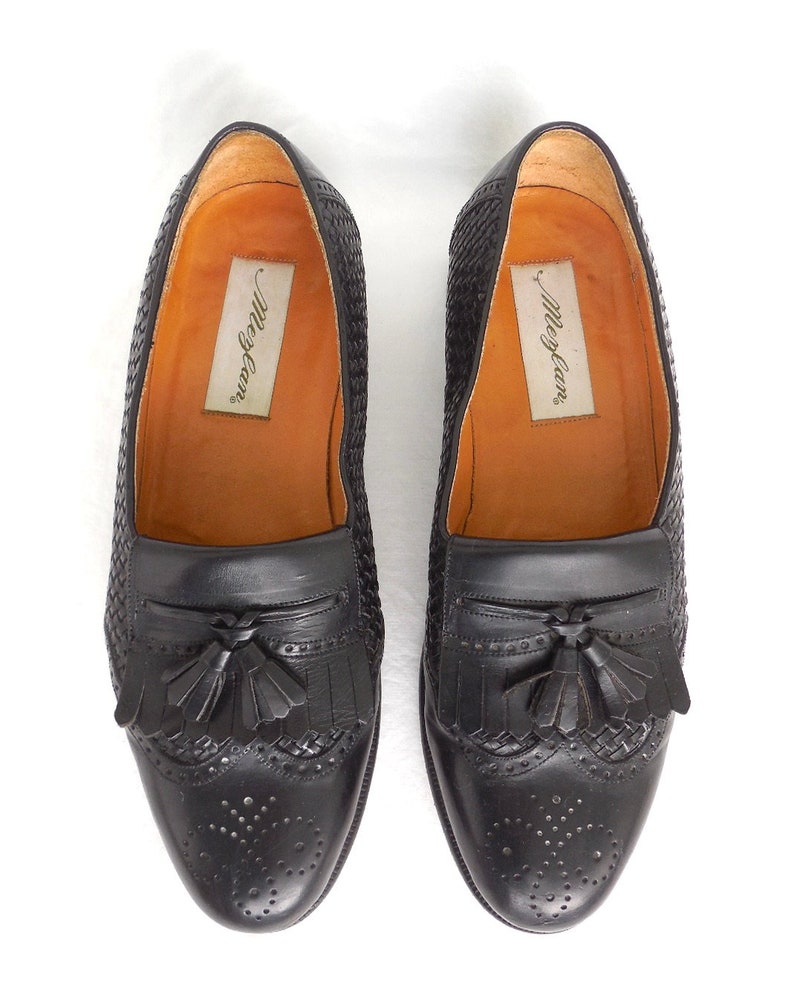 May include: A pair of black leather tassel loafers with a woven pattern and a perforated design. The loafers have the brand name "Merry-Go-Round" printed on the insole.