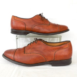 May include: A pair of brown leather dress shoes with a cap toe design. The shoes have dark brown laces and soles. The shoes are displayed on a clear glass block against a white background.