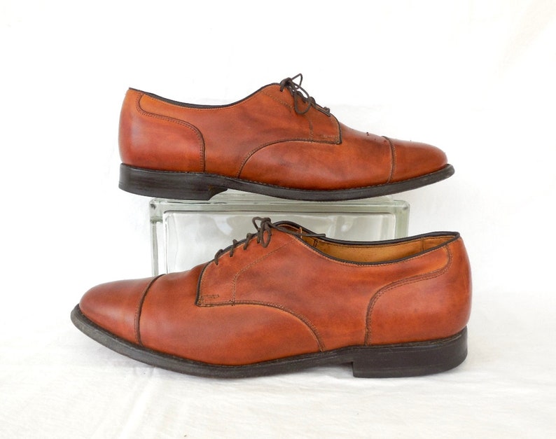 May include: A pair of brown leather dress shoes with a cap toe design. The shoes have dark brown laces and soles. The shoes are displayed on a clear glass block against a white background.