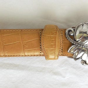 May include: Tan leather belt with a silver-coloured, ornate leaf-shaped buckle. The belt has a crocodile skin texture. The buckle is detailed with small, round embellishments. A stylish accessory for any outfit.